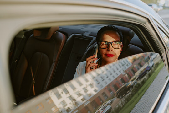Middle aged Caucasian woman sitting in back seat of car talking on smartphone, wearing glasses, looking out window with thoughtful expression, city street visible outside