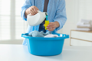 Woman pouring detergent into cap over plastic basin with towels at white table indoors, closeup