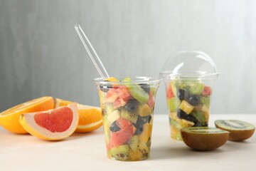 Tasty fruit salad in plastic cups and ingredients on beige table, closeup. Healthy snack