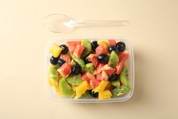 Tasty fruit salad in plastic container and spoon on beige background, flat lay. Healthy snack