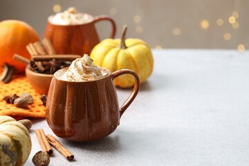 Tasty pumpkin latte with whipped cream in cups, spices and ingredients on light grey table against blurred lights, space for text. Bokeh effect