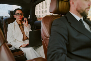 Caucasian middle aged woman sitting in back seat of car talking on phone and working on laptop while unrecognizable man driving, both wearing business attire, city buildings visible outside window