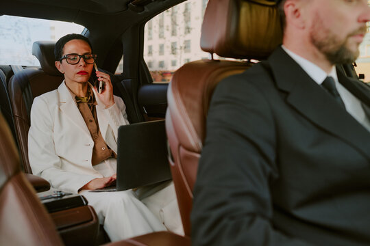 Caucasian middle aged woman sitting in back seat of car talking on phone and working on laptop while unrecognizable man driving, both wearing business attire, city buildings visible outside window