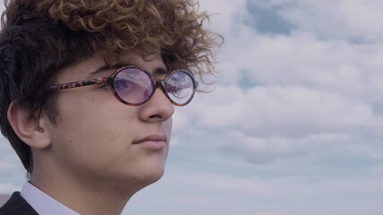 Portrait of a curly-haired teenager with glasses and a mature business suit against a backdrop of a hill and blue sky. The camera pans around.