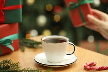 Woman holding gift box and wooden table with aromatic coffee, candy cane and fir tree branches against blurred lights, selective focus. Christmas greeting card