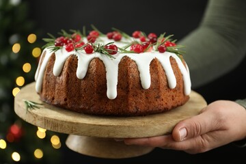 Woman holding delicious Christmas cake with icing, cranberries and rosemary against black background with blurred lights, closeup. Bokeh effect