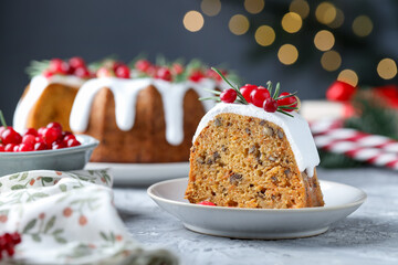 Piece of delicious Christmas cake with icing, cranberries and rosemary on grey textured table against blurred lights, closeup. Bokeh effect