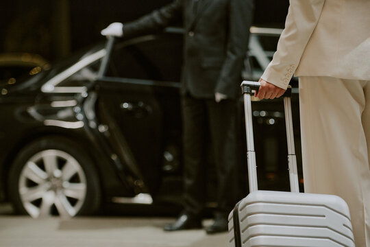 Caucasian middle aged man holding suitcase standing near luxury car while chauffeur in uniform opening door, business travel concept, partial body view, no faces visible