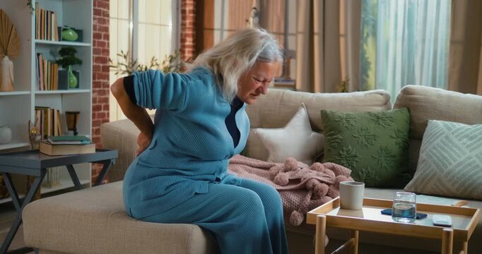 Middle-aged woman suffering from severe lower back pain, grimacing and rubbing her back while struggling to stand up from a sofa. Concept of discomfort, tension, and physical strain.