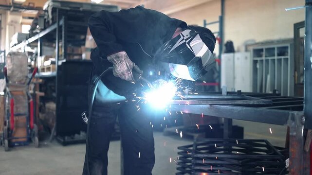 Industrial worker wearing a protective mask and gloves welding a metal structure in a workshop, creating bright flashes of light and sparks from the high-temperature arc process