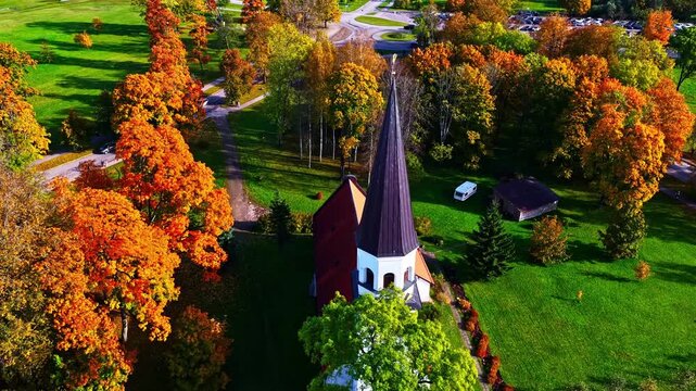 A circling pan shot orbits a white church with black steeple amid fiery fall foliage, revealing paths and rural serenity from above.