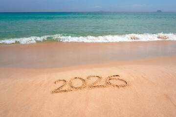 Soft golden sand features the number 2026 drawn neatly, while gentle ocean waves lap against the shore under clear blue sky on a sunny beach day