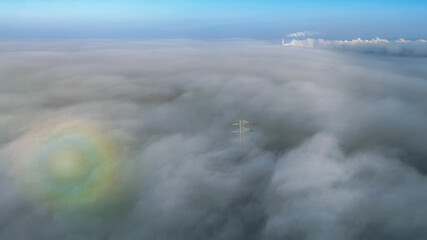 High-voltage tower emerging from dense fog. Aerial view with atmospheric clouds and subtle rainbow phenomenon.