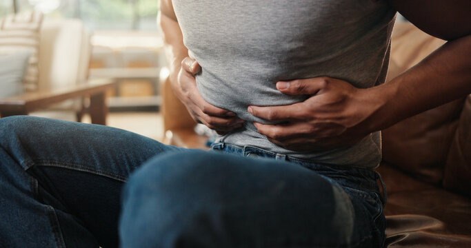 Hands, ibs and tummy with man on sofa in living room of home for indigestion symptoms closeup. Abdomen, pain and stomach ache with person in apartment for chronic digestive or gastro condition