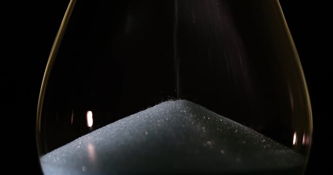 Glitter sand falls from top chamber of hourglass on black background. Grains poured slowly at constant rate creating continuous flow of time