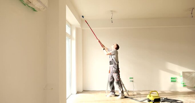 Man in uniform paints ceiling in white color with roller brush at home. Builder maintains interior design style with light pigment in apartment
