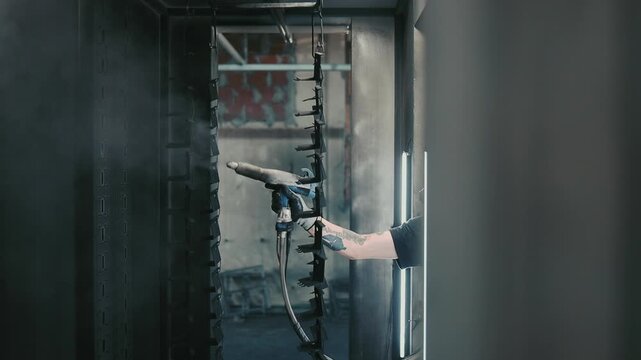 Professional male worker with a respirator mask using a spray gun to apply powder coating on metal components hanging from an overhead conveyor line in an industrial spray booth
