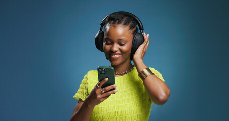 Headphones, phone and black woman in studio with music, radio or song for rhythm and energy. Happy, audio tech and African person listening to album on cellphone with mobile app by blue background. © peopleimages.com