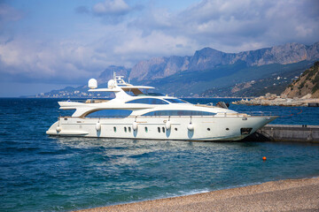 A luxurious white expensive yacht stands on a pier in the port of the sea against the backdrop of picturesque mountains. The concept of expensive luxury living