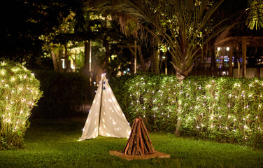 Enchanting garden evening with a cozy white teepee tent, glowing string lights, and a rustic campfire setup on lush grass. © Pixel World