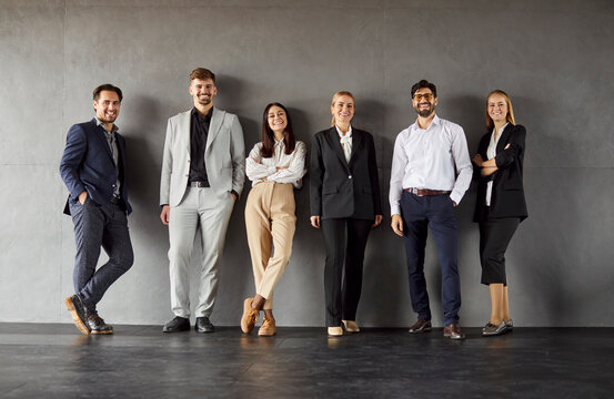 Group full length portrait of confident business professionals standing against plain wall, smiling and looking at camera. Diverse team in formal and smart casual attire, showing unity and positivity.