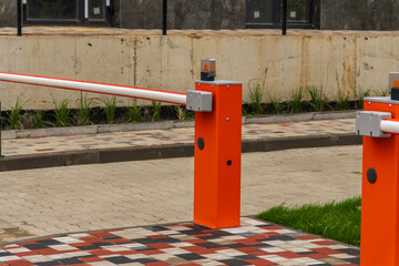 Bright orange barrier poles secure the entrance to a busy urban parking lot surrounded by greenery and pavement on a cloudy day