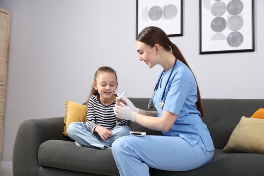 Diabetes. Smiling doctor in medical gloves checking little girl's blood sugar level with lancet pen on sofa indoors - Powered by Adobe