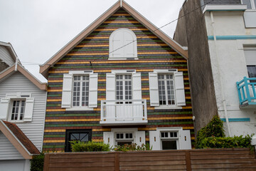 Façade avec fenêtres et petit balcon blancs d'une maison moderne, recouverte d'un bardage verni de couleurs brune, jaune et verte.