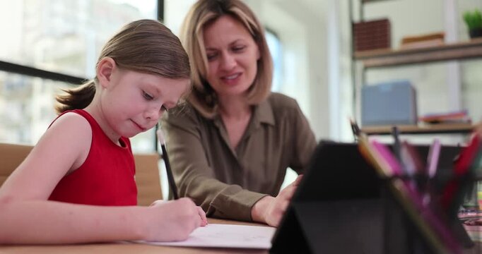 Mother and daughter sit together at table and do homework. Mom carefully explains grammar rule to schoolgirl and child writes in notebook. Slow motion