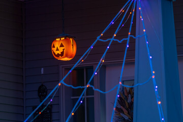 Glowing jack-o'-lantern with blue string spider web, orange and purple lights and skeleton on a porch at dusk for Halloween.