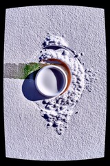 A white container is tipped, spilling white powder onto a textured surface, creating abstract shapes and shadows.