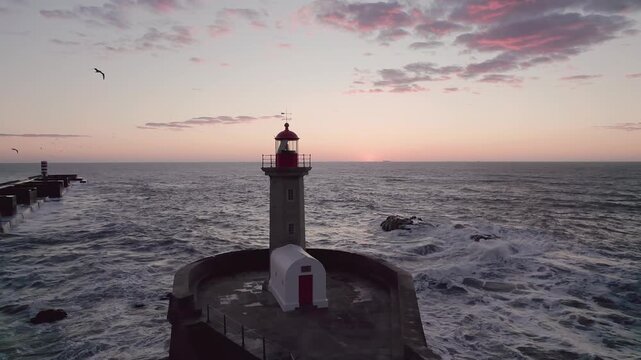 Red-and-white striped Farol da Barra lighthouse tower stands on rocky breakwater with beacon lit during golden hour dusk, Atlantic waters and horizon sky glowing amber.