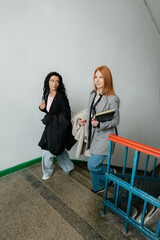 Two women walk up stairs, chatting and carrying coats and folders in a casual business setting.