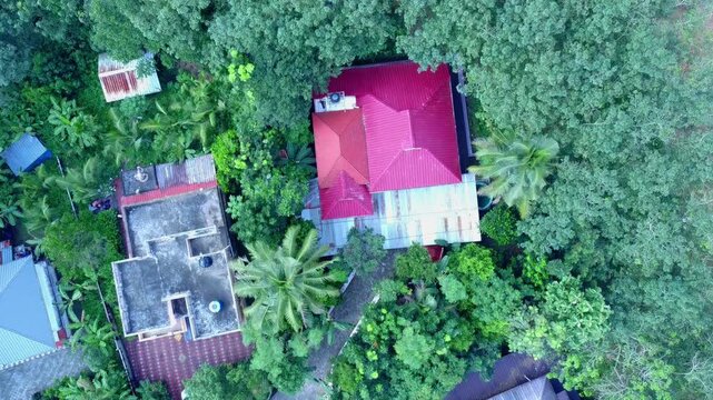 Aerial zoom on a vibrant red-roofed house in a dense tropical residential area, next to a newly marked land plot for construction and real estate development in Kerala.