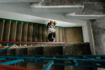 Two friends on a stair landing read a book, surrounded by blue railings in a quiet interior.