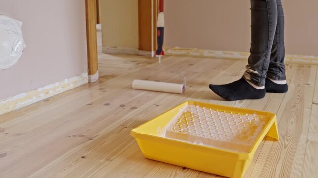 A person stands on a hardwood floor, using a long red-handled roller to apply varnish from a yellow tray near a taped baseboard.