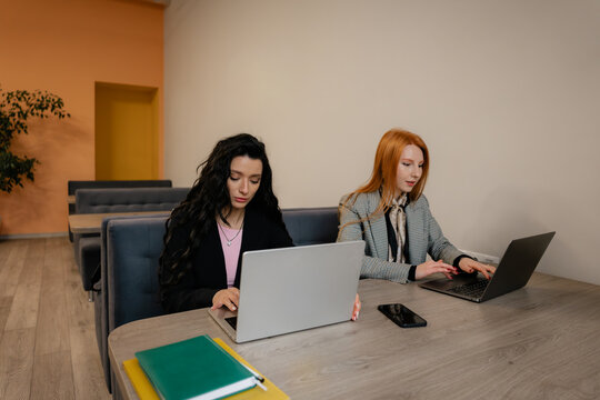 Two women work on laptops at a bright, modern office table, showcasing collaboration and productivity.