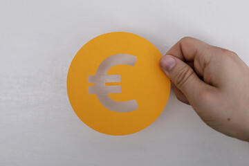 A hand holds a yellow cutout of the Euro currency symbol against a textured white background....