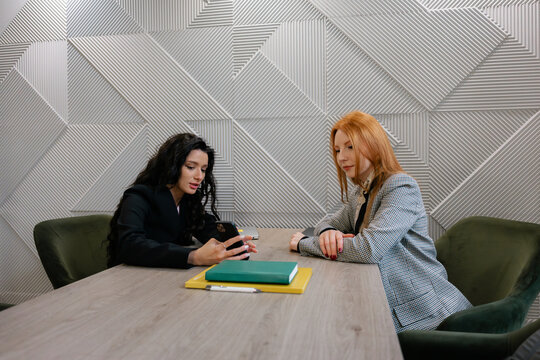 Two professional women sit at a large wood table, reviewing notes and sharing ideas.