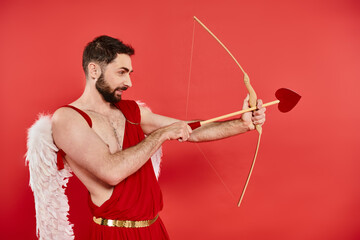 side view of bearded man in cupid costume aiming and archering with hear-shaped arrow on red