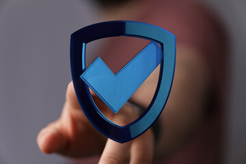 A hand interacts with a blue digital shield icon featuring a checkmark, symbolizing security, safety, and validation in the digital r
