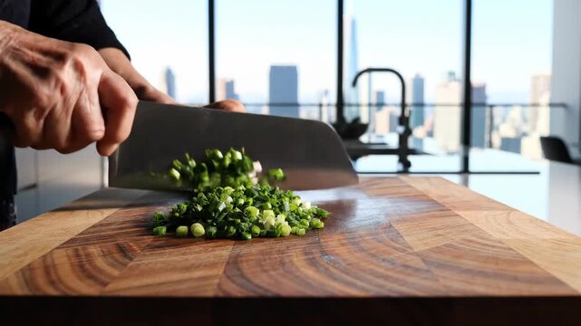 Hands using a large cleaver knife to chop fresh green onions on a wooden cutting board, preparing ingredients in a modern kitchen with a city skyline view