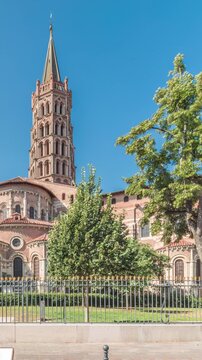 The cobbled parvis of the Basilica of Saint Sernin in Toulouse, France, timelapse hyperlapse. Largest romanesque church in Europe with brick bell tower, arched doors, green trees and blue sky