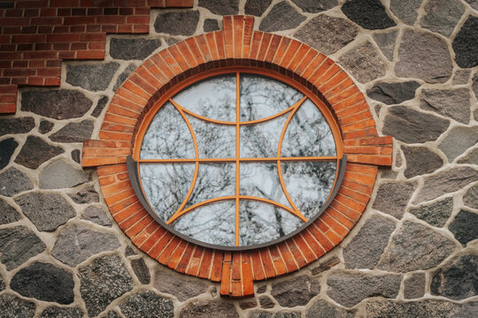 A round window with an ornate orange lattice design set into a red brick and natural stone wall, reflecting leafless trees in the glass. Architectural details with symmetry and texture.