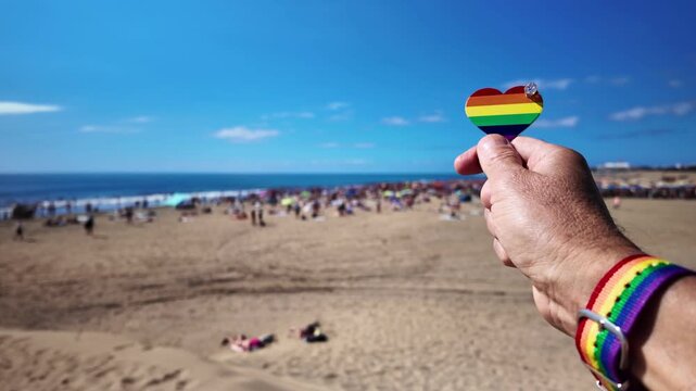 First-person view of a hand with a rainbow wristband holding a heart with the gay pride flag colors, symbolizing love and diversity at a sunny beach festival in Maspalomas