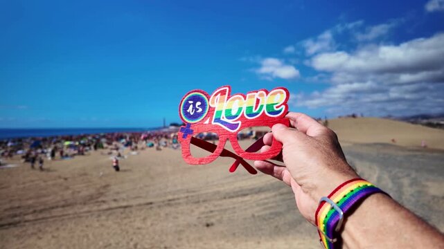 Man's hand wearing a rainbow flag bracelet holding a paper glasses with the inscription love is love at the winter gay pride beach party in Maspalomas under a clear blue sky