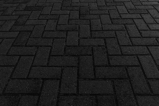 A close-up of dark brick paving with a "herringbone" pattern at night with minimal lighting creating subtle texture and shadows.