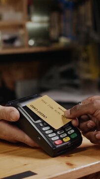 Vertical closeup of hand of unrecognizable customer paying for purchase with credit card, tapping it on terminal cashier holding
