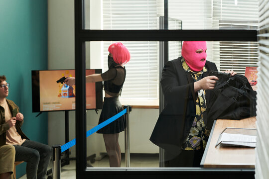 Two masked young adults, one Caucasian man wearing pink balaclava putting bag on counter, one woman with pink wig pointing handgun at seated Caucasian man in modern bank setting