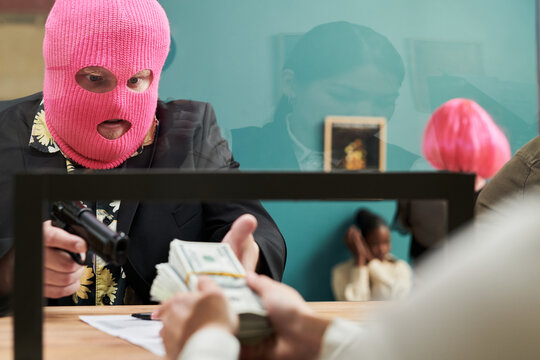 Caucasian man wearing pink ski mask pointing handgun at bank teller while receiving stack of cash, young Black girl sitting against wall covering ears